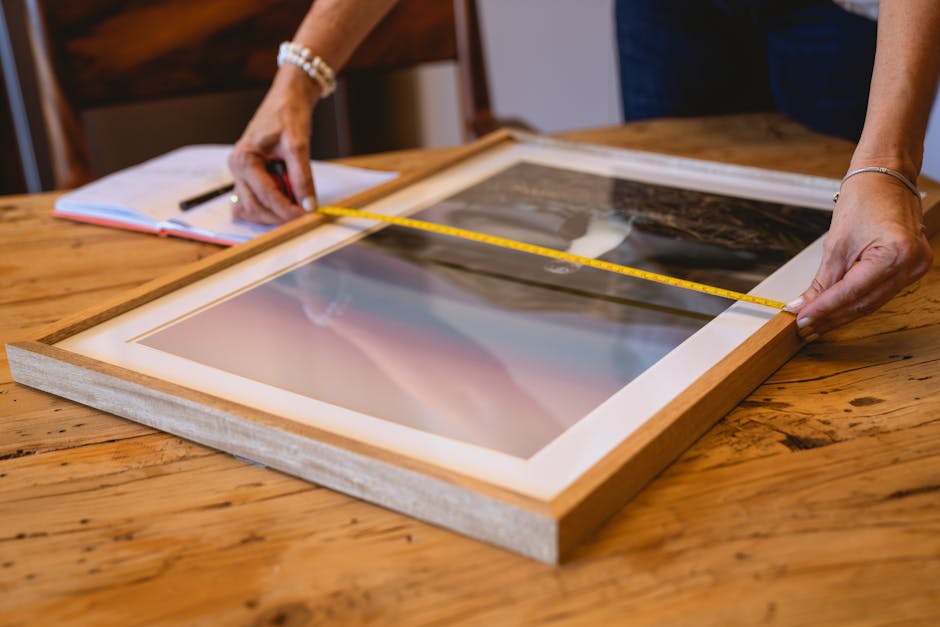 Hands using a tape measure to size a wooden picture frame on a wooden table indoors.